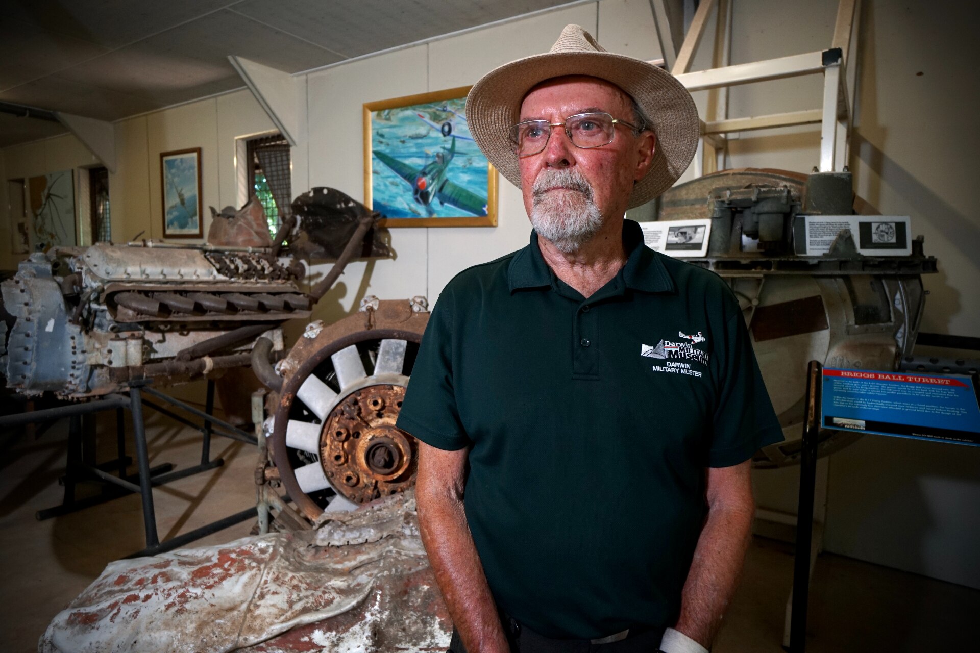 A man in a brimmed hat, glasses and black t-shirt standing inside a military museum.