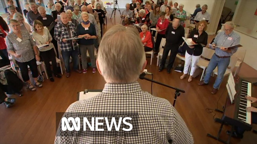 The back of a conductor's head can be seen in while he stands on stage in front of dozens of elderly people singing.