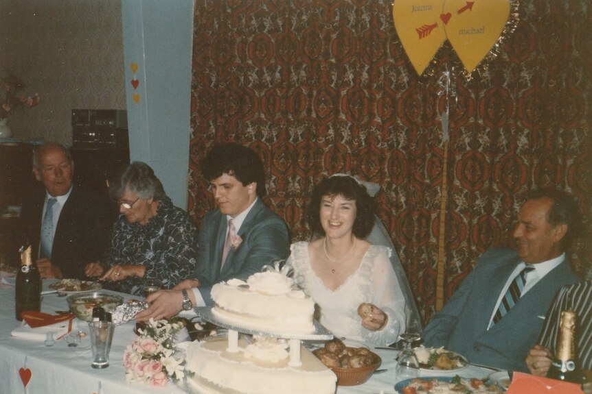 A bride and groom sitting at a table at their wedding, with their cake in front of them.