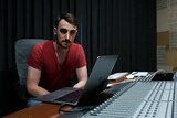 Sydney music producer George Tulloch wears a red t-shirt sitting in front of a music console with an open lap top