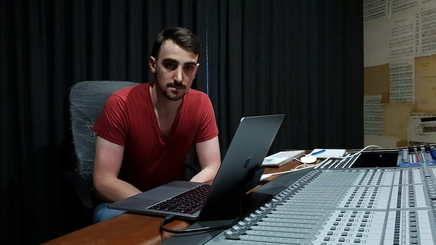 Sydney music producer George Tulloch wears a red t-shirt sitting in front of a music console with an open lap top