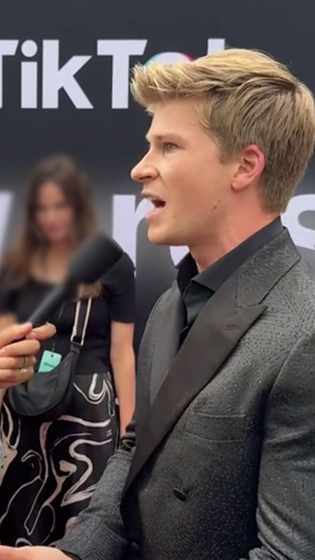 A young blond man speaks into a microphone on a red carpet. He wears a black suit. 