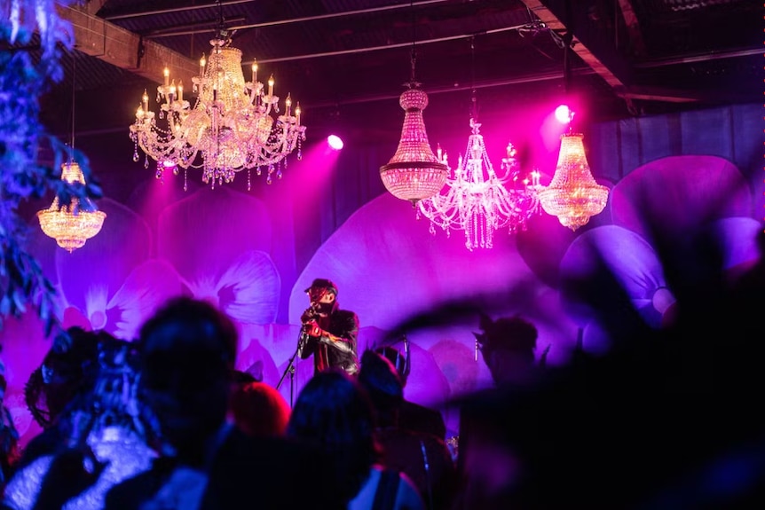chandeliers hang from the ceiling. A man is playing on stage and the lights are purple and pink.