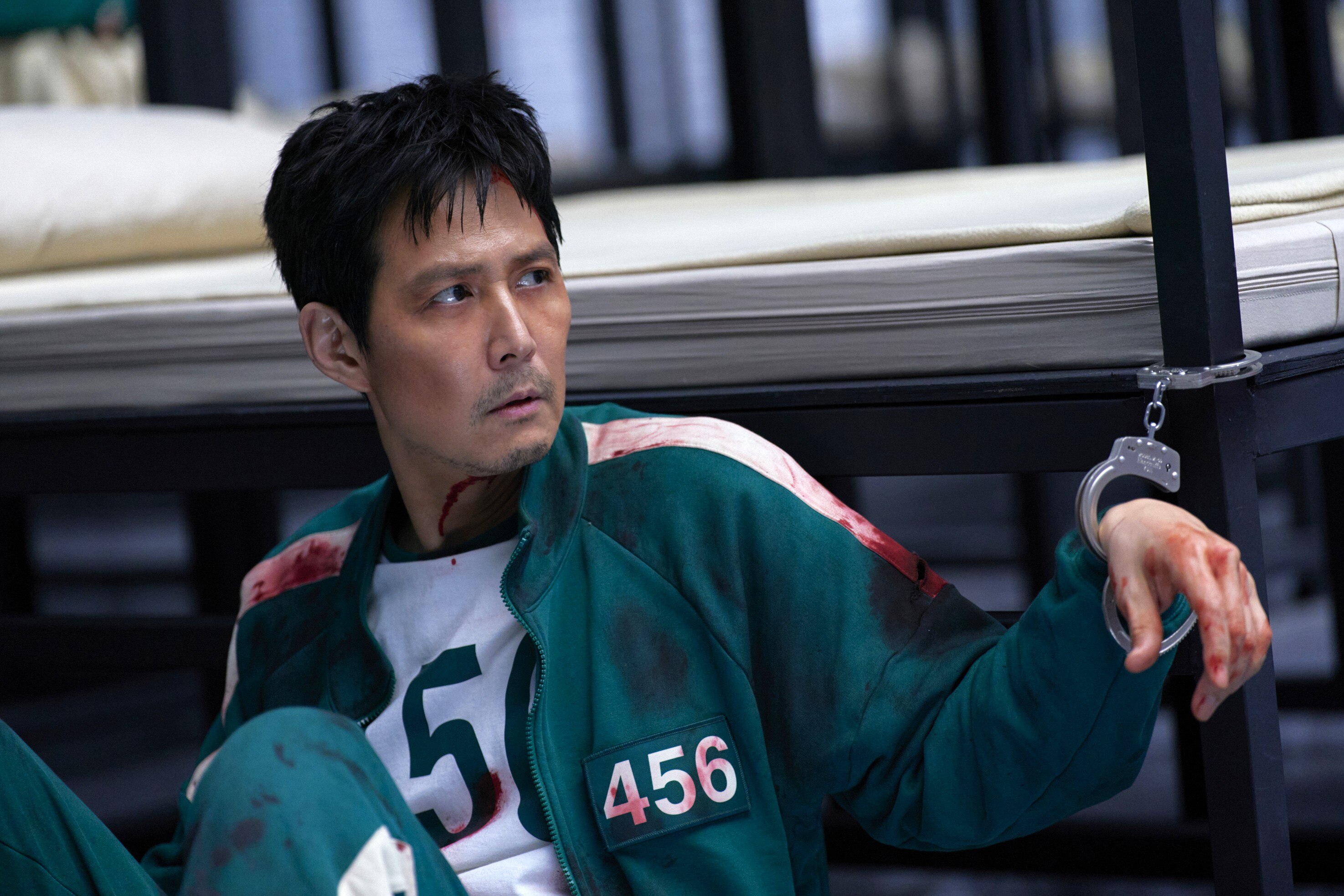 A man in a bloody green tracksuit sits handcuffed to a bed