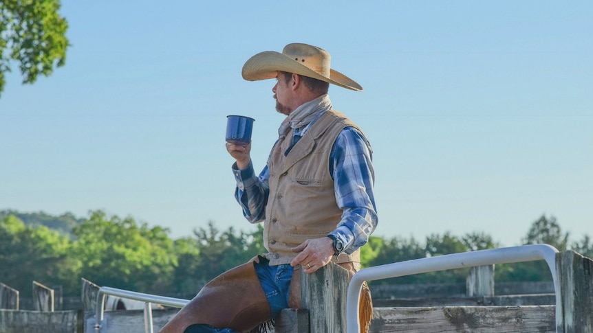 On a sunny day, a cowboy holding a mug straddles a fence and looks behind himself.