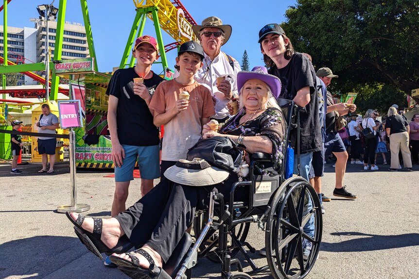In front of a rollercoaster poses an elderly woman in a wheelchair, surrounded by an elderly man and three teenage boys.