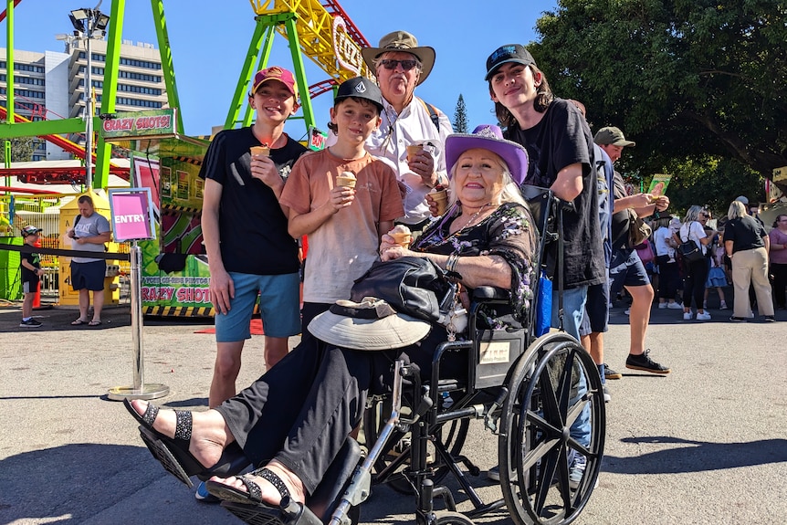 In front of a rollercoaster poses an elderly woman in a wheelchair, surrounded by an elderly man and three teenage boys.