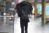 A man in black carrying a backpack walks down a city street in the rain holding an umbrella.
