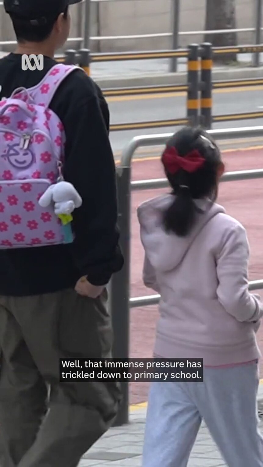 An adult with black hair and a pink backpack walks with a child on an urban street