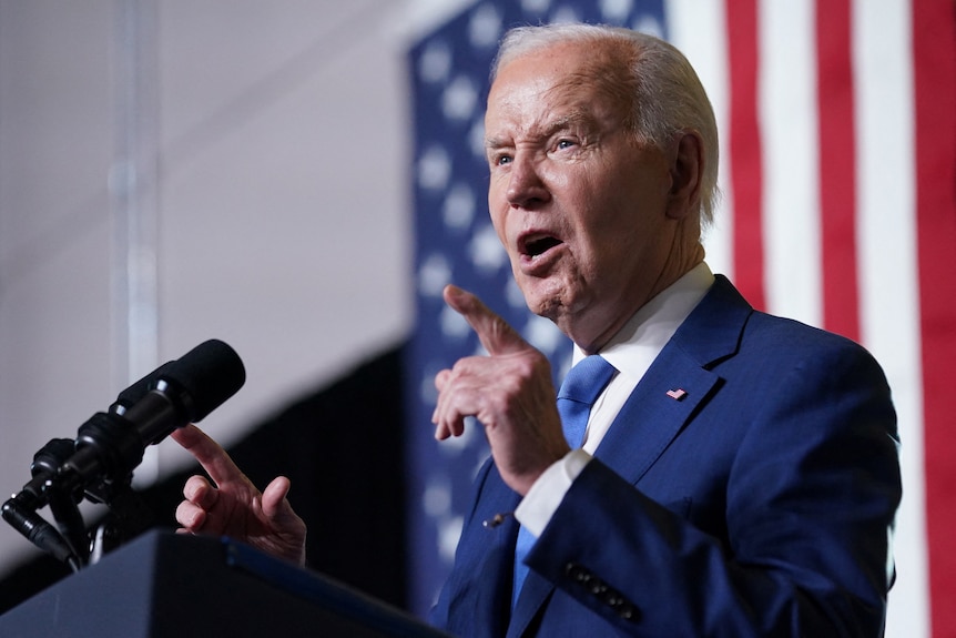 Joe Biden speaks at a microphone with an American flag behind him.