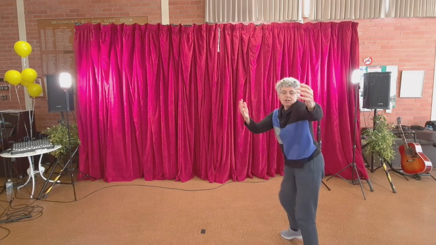 An ageing woman dances expressively against a red curtain on a stage.