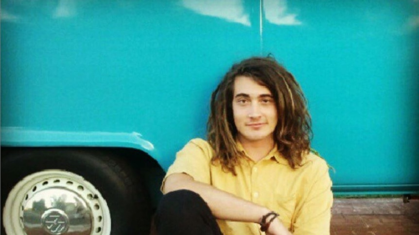 Tristan Naudi sitting in front of his aquamarine Kombi van, he is wearing a bright yellow shirt.