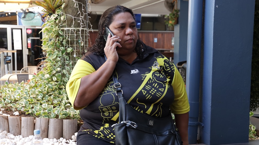 An Indigenous woman sits on a bench talking on the phone.