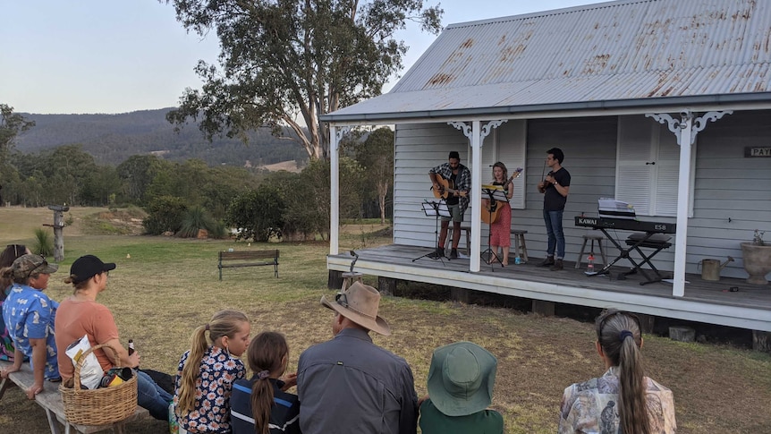 Three performers sing and play instruments on the patio of a country house, while a group watch on as an audience.