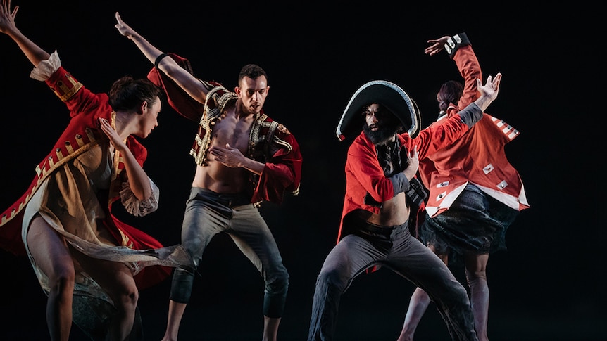 Four male dancers wear red and black colonial style costumes perform on stage with arms raised.