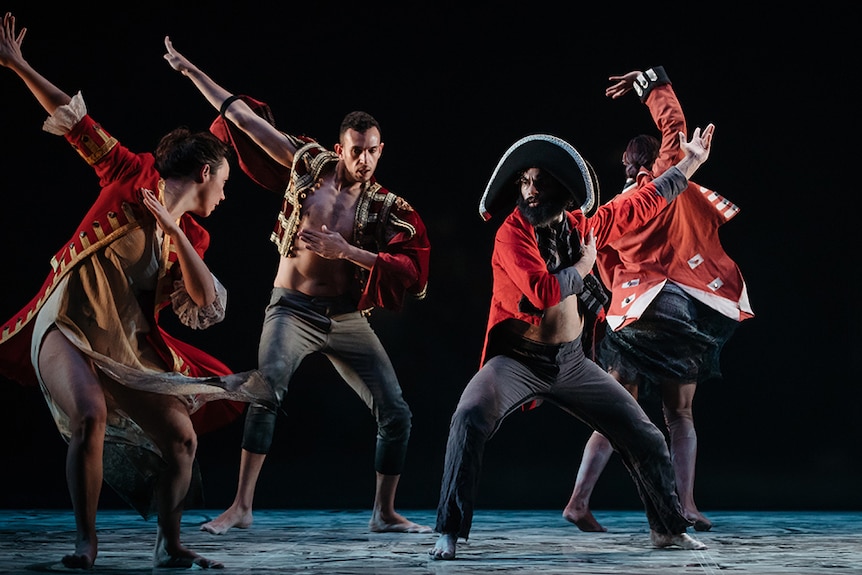 Four male dancers wear red and black colonial style costumes perform on stage with arms raised.