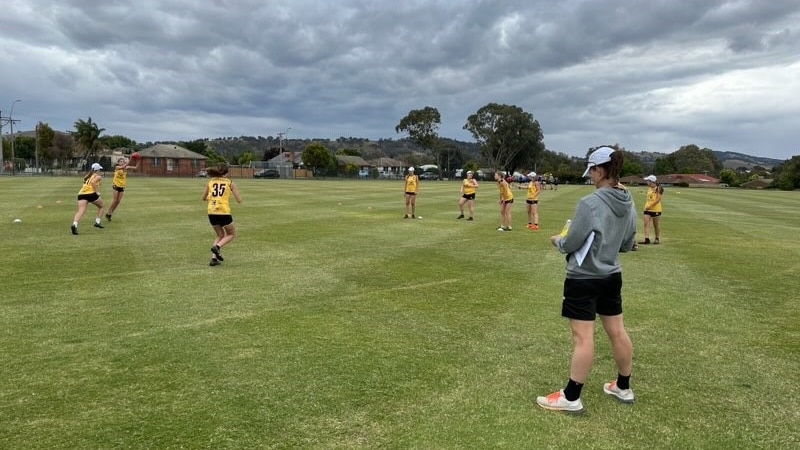 AFLW coach Emma Mackie stands on footy oval watching young players train.