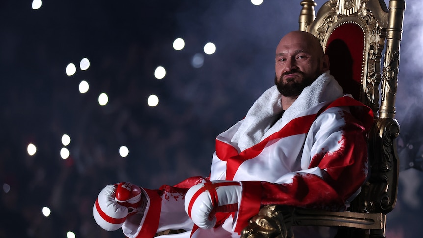 A bald man wearing a robe in English flag colours and with gloves on his hands sits on a throne.