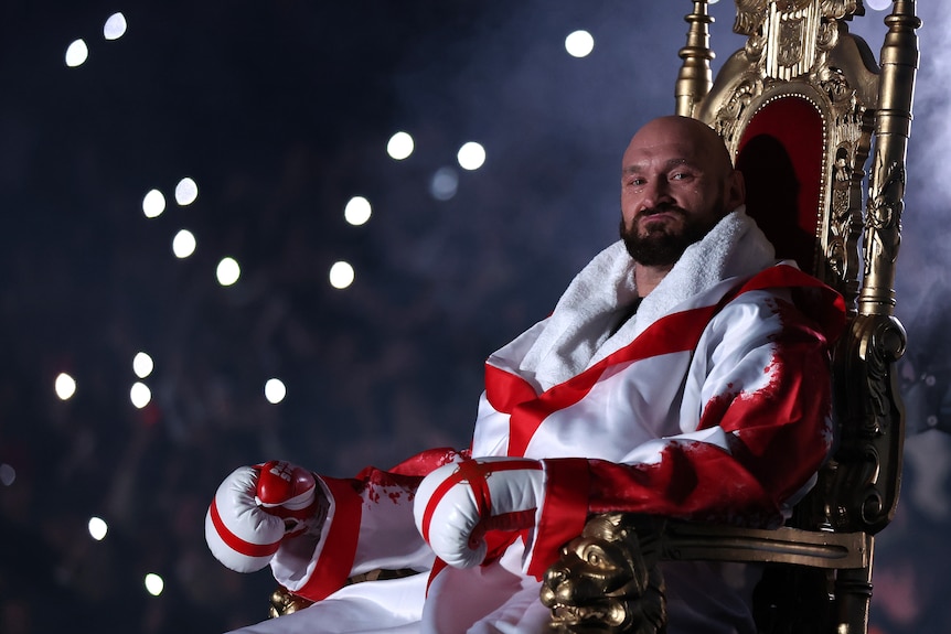 A bald man wearing a robe in English flag colours and with gloves on his hands sits on a throne.
