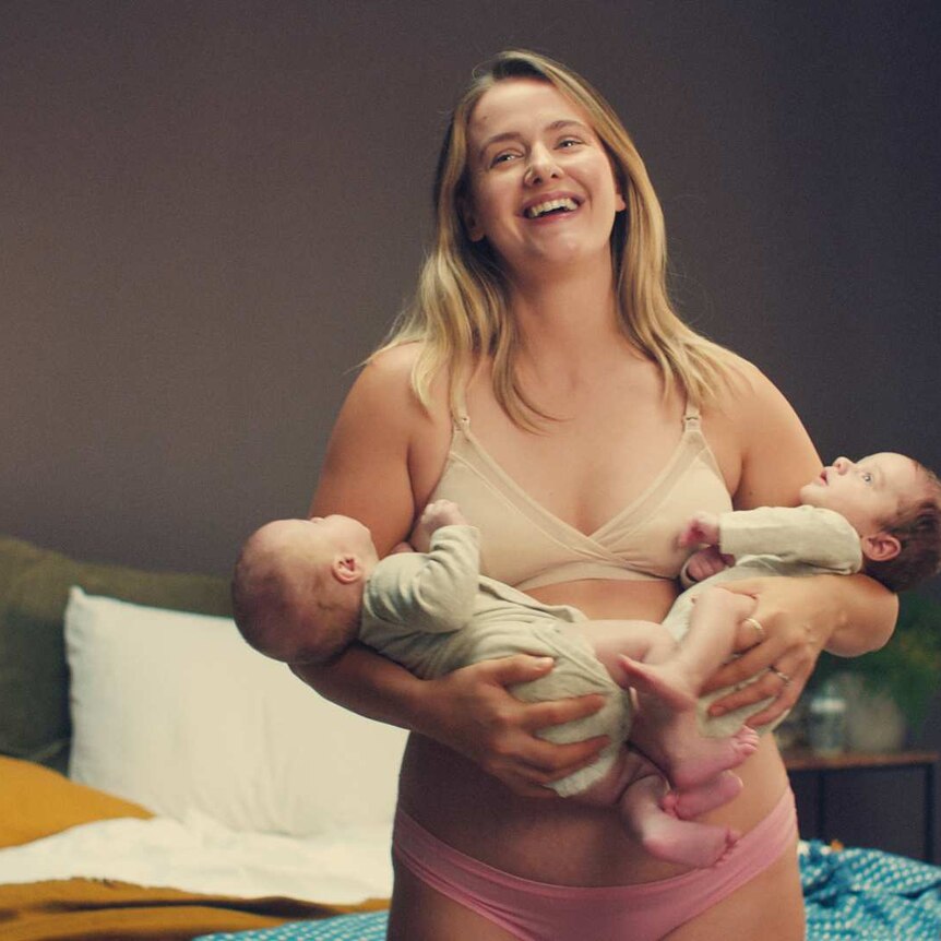 a woman stands in her underwear holding two young babies possibly twins, she looks laughingly at someone in the distance