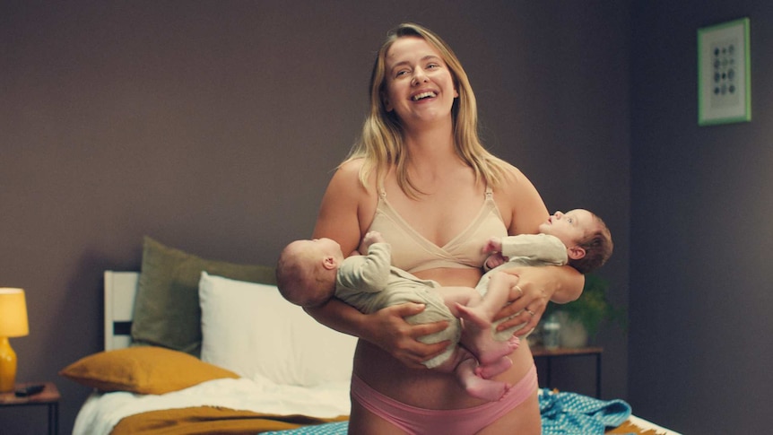 a woman stands in her underwear holding two young babies possibly twins, she looks laughingly at someone in the distance