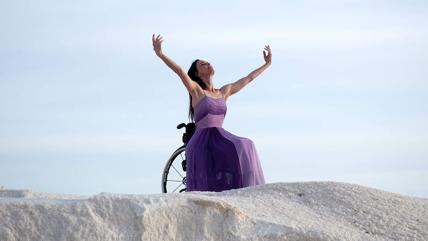 A white woman with brown hair, wearing a purple dress and seated in a wheelchair, dances with her arms in the air.