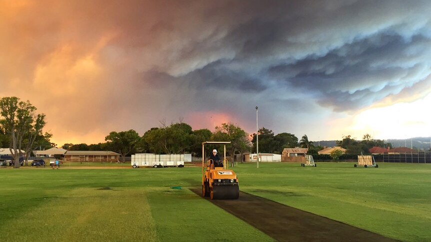 A curator in a roller works on a cricket pitch as a large menacing bushfire ash cloud looms overhead.