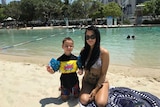 A tatooed woman in swimsuit and sunglasses sits next to small boy in floaties on sunny city beach.