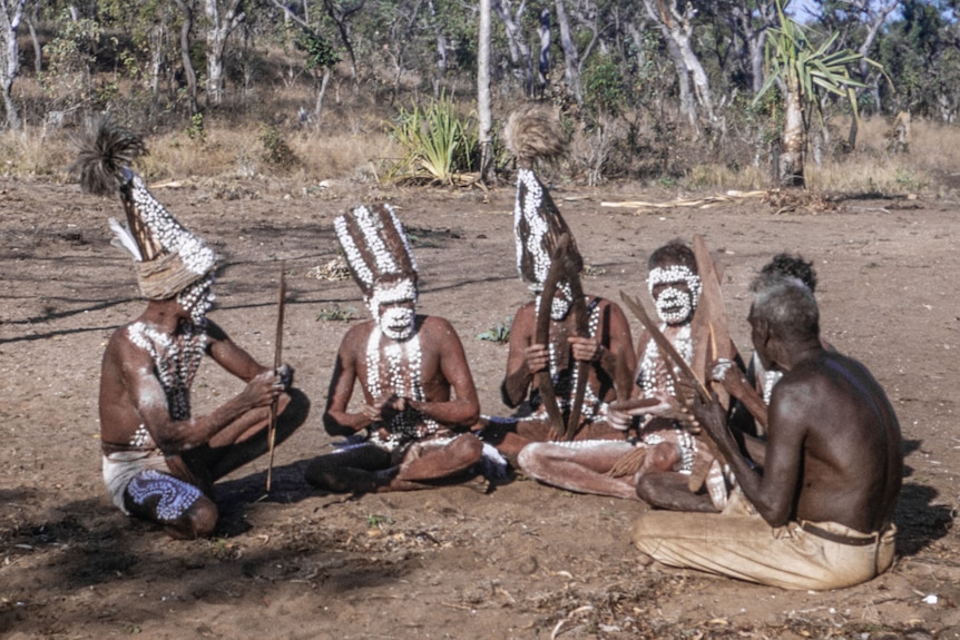 Six Indigenous men sit on the ground in ceremonial paint and headdresses.