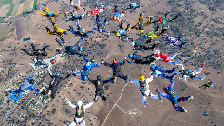 An assortment of skydivers in a circlular formation, linked by arms and legs, free-fall towards the dry ground below.