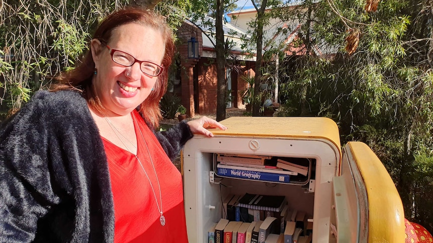 A woman stands beside an old bar fridge, which has been repurposed as a bookshelf.