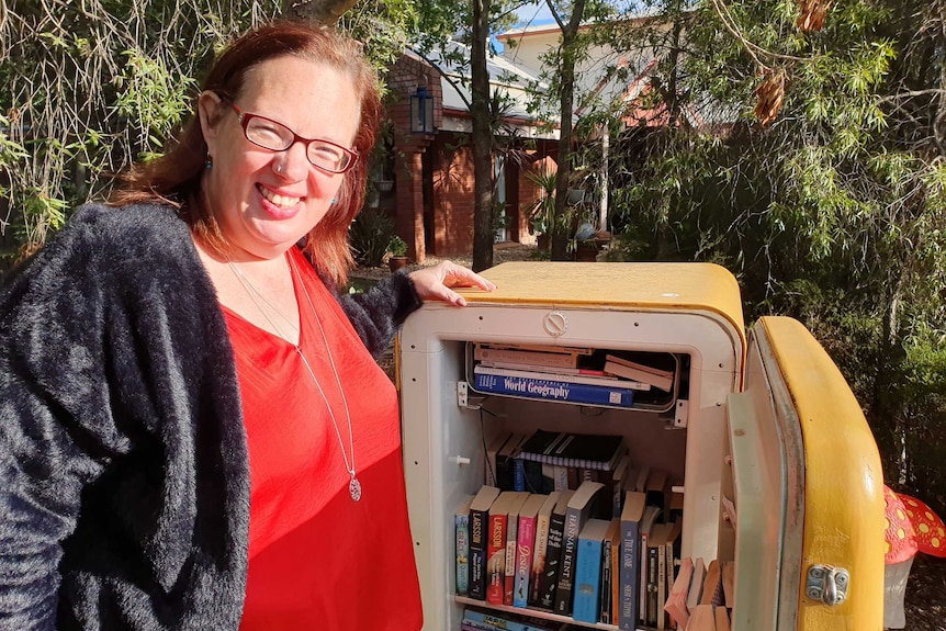 A woman stands beside an old bar fridge, which has been repurposed as a bookshelf.