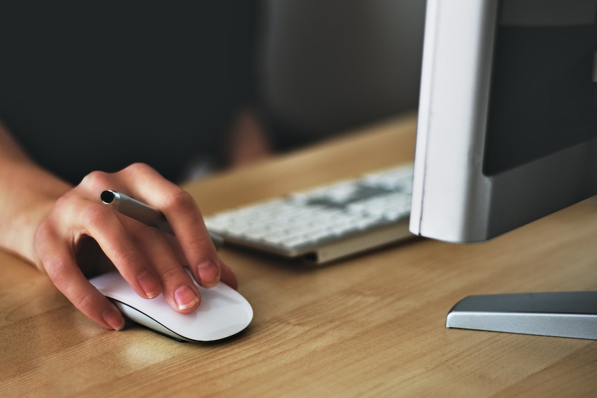 A hand on a mouse with a side of the monitor shown and the keyboard in the background