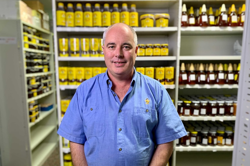 A man stands looking directly at the camera in front of shelves of jars of honey.