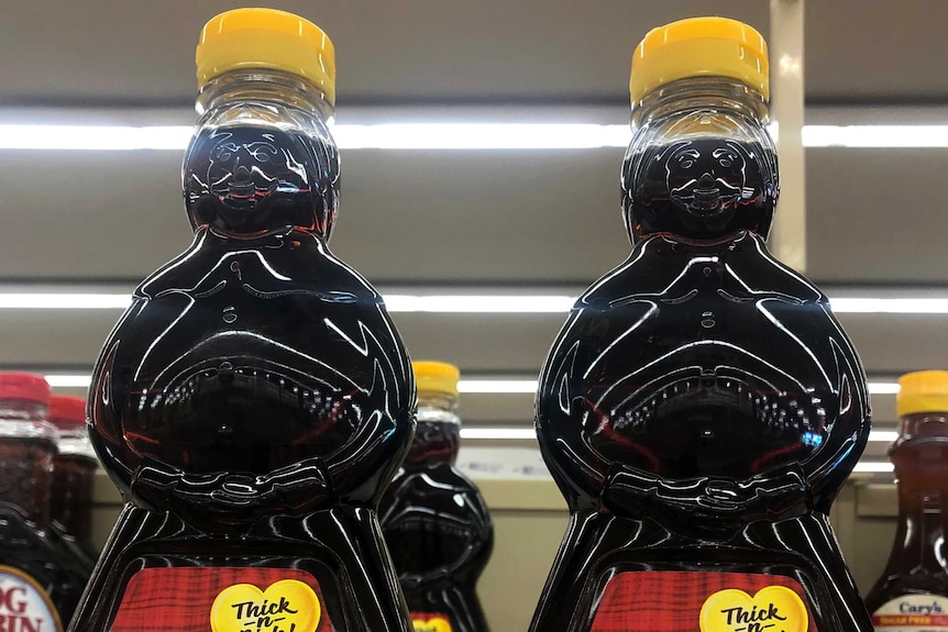 Two clear bottles filled with syrup are seen on a shelf with more behind them. They are in the shape of two black women.
