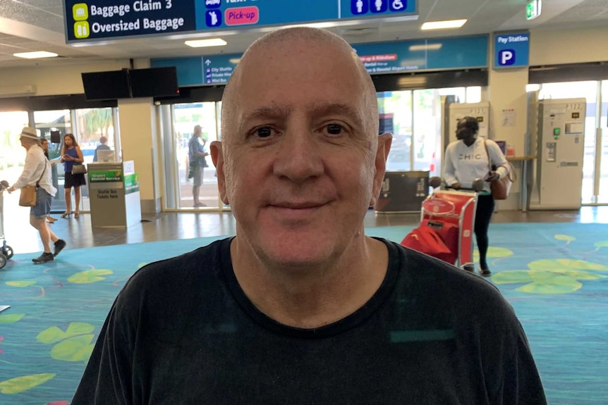 Jeff Toll is inside Darwin Airport. He is wearing a black t-shirt and looks happy.