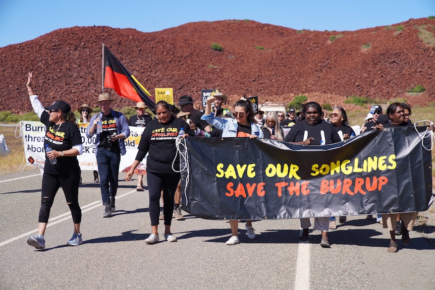 A group of people walk along an outback road carrying signs and an Aboriginal flag.