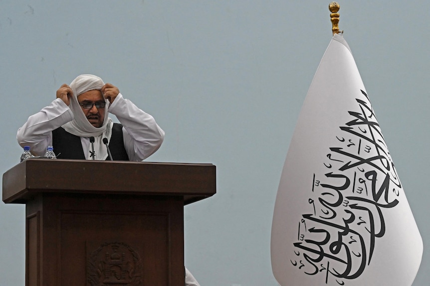 Abdul Baqi Haqqani holds his hands to his head as he stands near a Taliban flag.