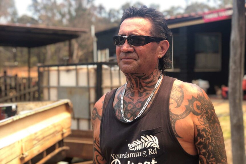 A man in a singlet and tattoos stands on his rural property