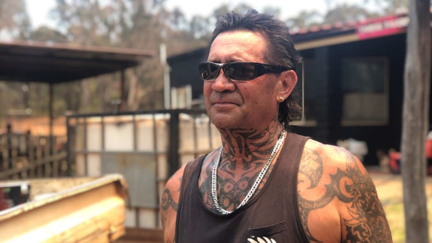 A man in a singlet and tattoos stands on his rural property