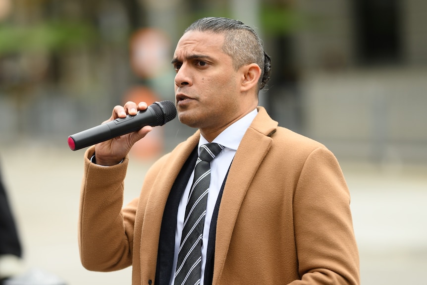 Dr Aziz wears a cream coat over a grey suit and white shirt and holds a microphone and a RACGP sign