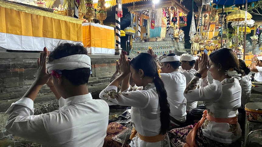 Men and women in traditional Balinese dress hold their hands to their heads in prayer at an outdoor temple in Bali