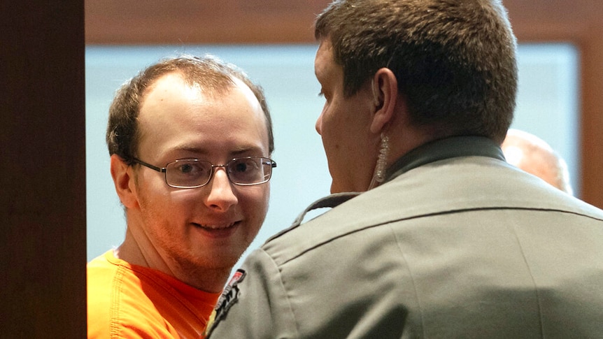 A man wearing an orange prison uniform and glasses smiles, looking towards the direction of the camera.