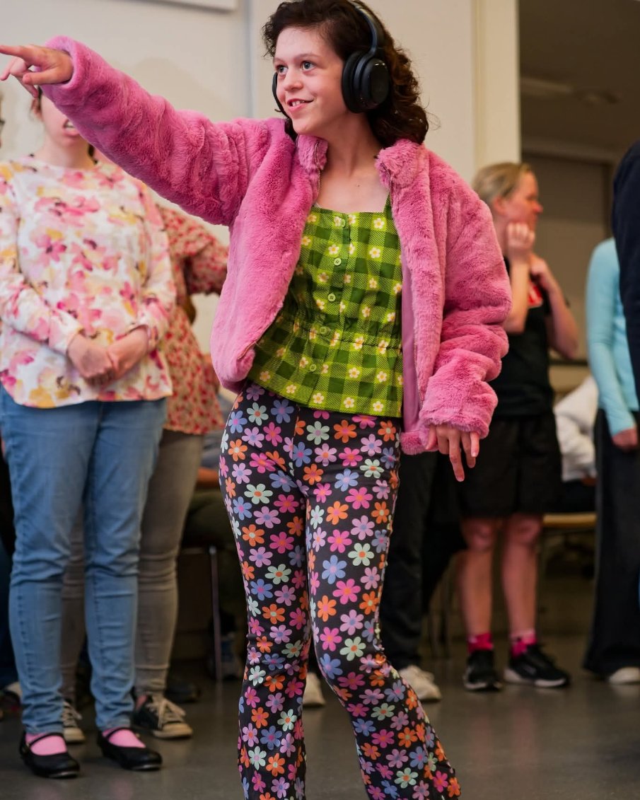 A teenage girl dancing. She is wearing headphones, a pink jacket and green top.