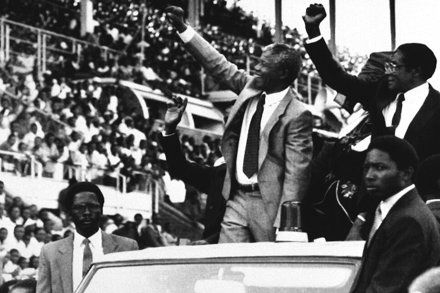 A black and white photo shows Nelson Mandela and Robert Mugabe with raised fists as they greet crowds in an open-top car.