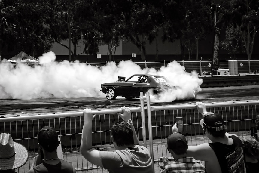 spectators watch a car doing burnouts from behind a metal fence and barricades
