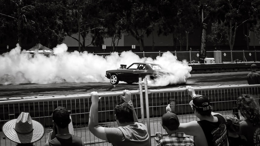 A black and white photo of spectators watching from behind a metal fence and barricades a car doing burnouts.