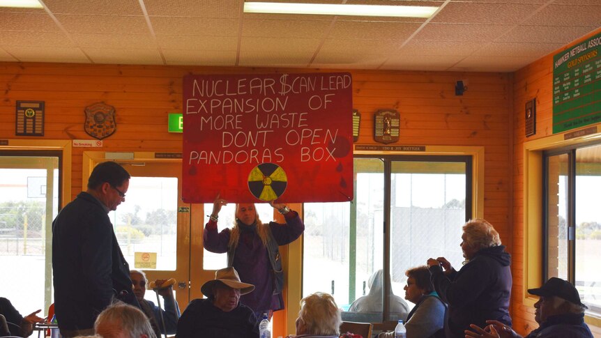 A man holds up a red sign protesting nuclear waste storage, community members look on.