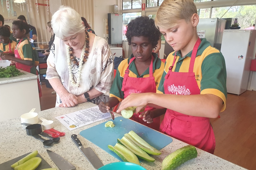 Two male students at kitchen bench slicing cucumbers with Stephanei Alexander watching on