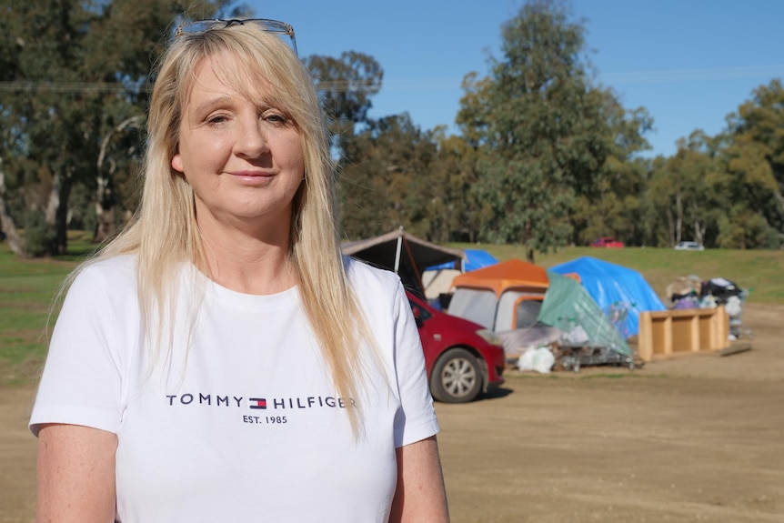 A woman with a fringe and long hair stands in front of tents and shopping trolleys.